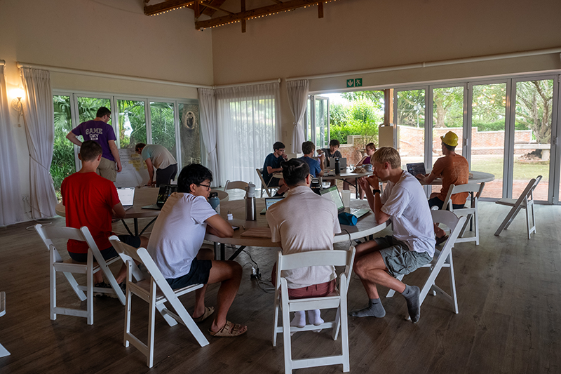Group of students working on laptops and talking at tables in a bright room with large windows and an open glass door to a garden.