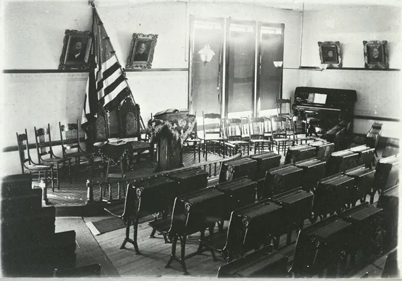 Historical interior of a meeting hall featuring rows of benches, a podium at the front, and an American flag.