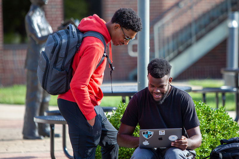 A student sitting outside showing another student something on his laptop