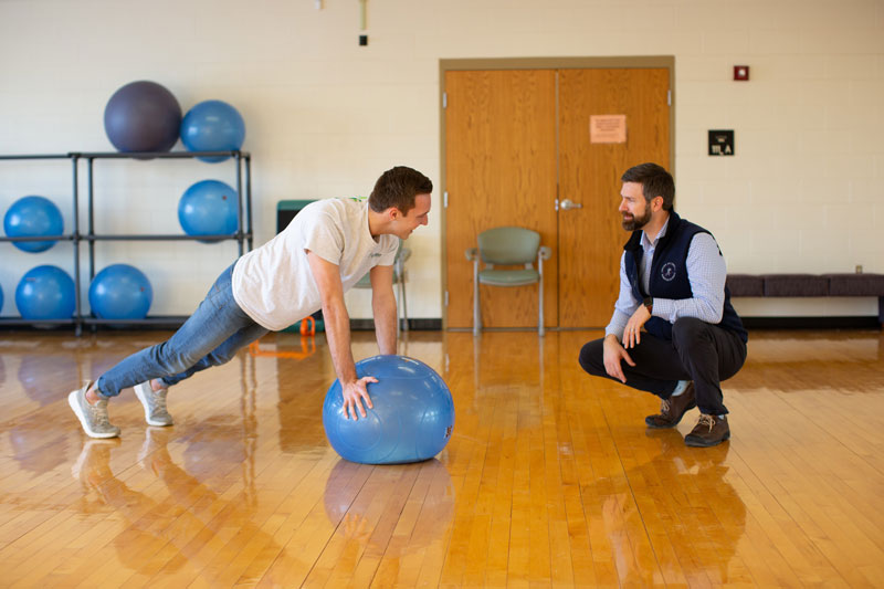 A professor watching a student do a push-up on an exercise ball