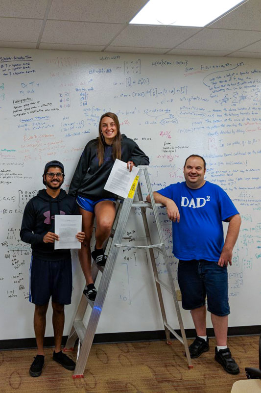 Two students and a professor standing in front of a whiteboard wall