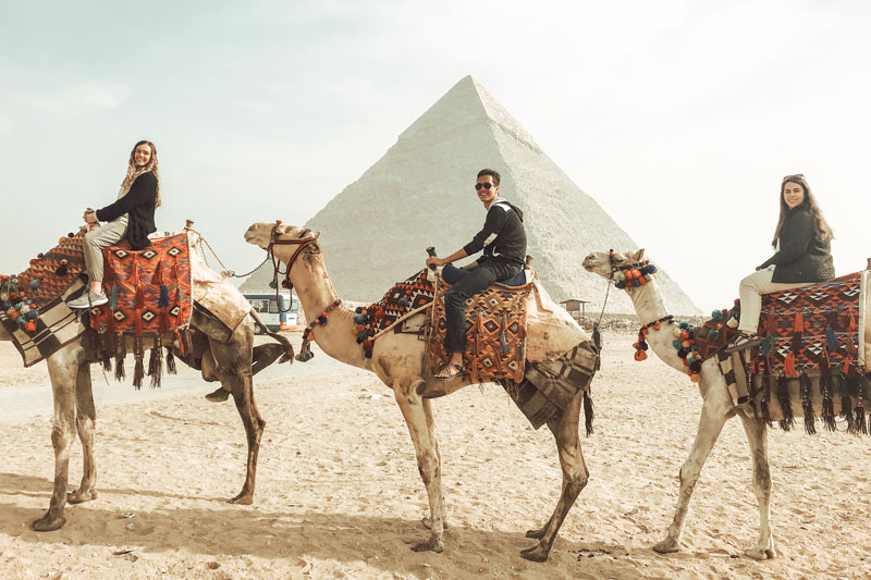 Three students riding camels in front of the Pyramids of Giza