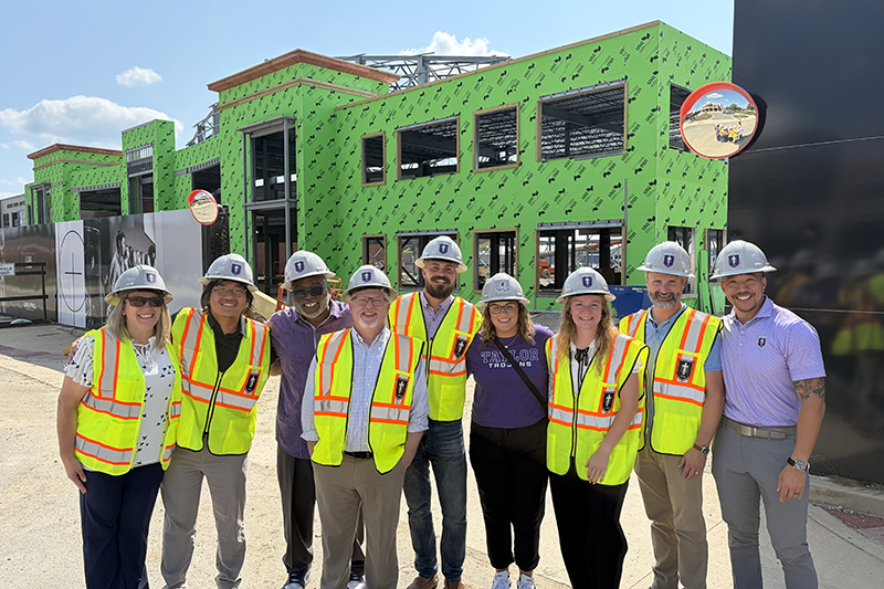 Group of people wearing hard hats and high-visibility safety vests pose in front of a green-sheathed building under construction.