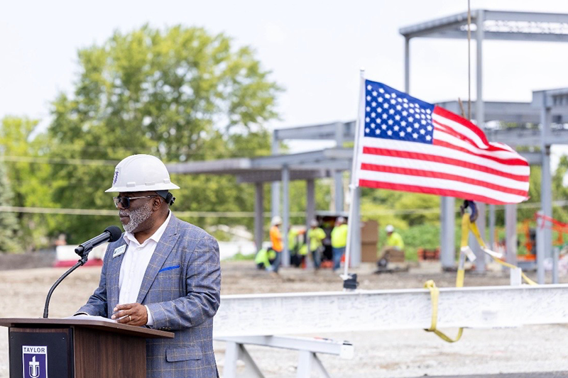 Speaker in a hard hat talks at a podium at a construction site, with an American flag and workers in the background.