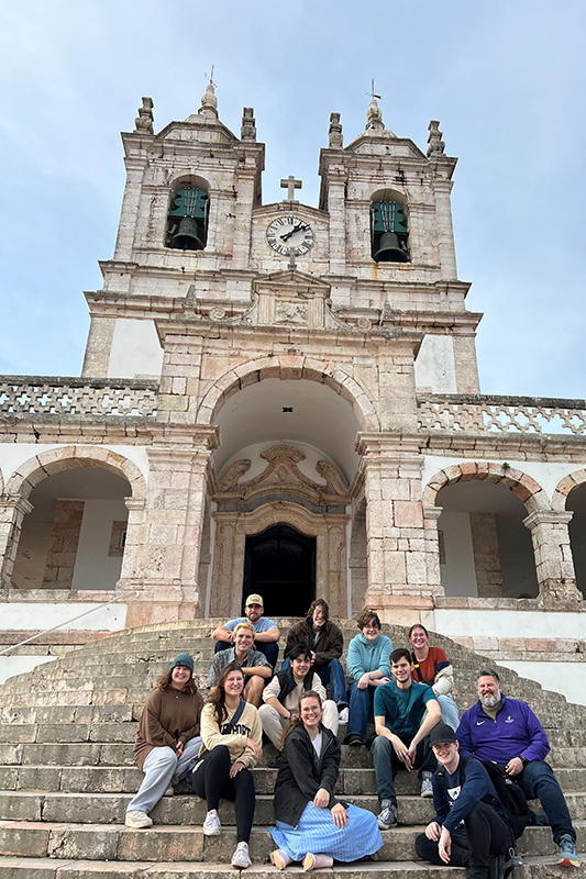 Group of people sitting on stone steps in front of a historic building with twin bell towers and a central clock.