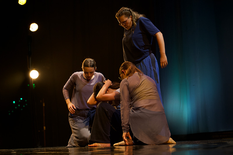 Four dancers in blue and gray costumes cluster around a seated performer on a dimly lit stage with spotlights.
