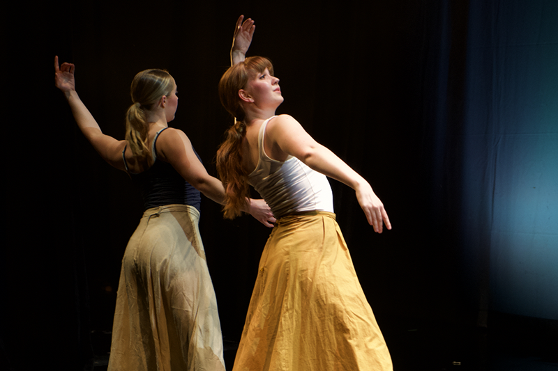 Two dancers on a dark stage pose mid-performance with arms raised, wearing long skirts under warm spotlight.