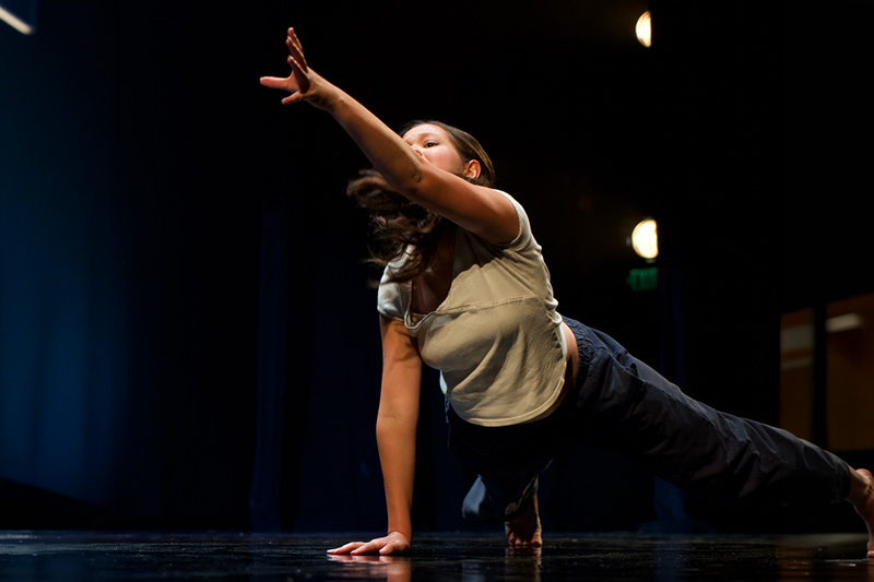 Contemporary dancer holds a dramatic floor pose, supporting herself with one hand while reaching high into the stage lights.