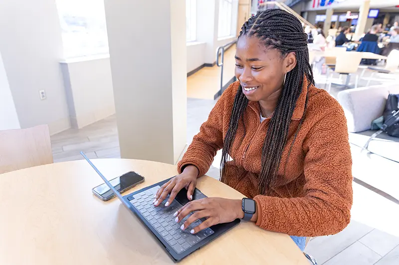 Student with braided hair smiles while typing on a laptop at a table in a bright campus common area.