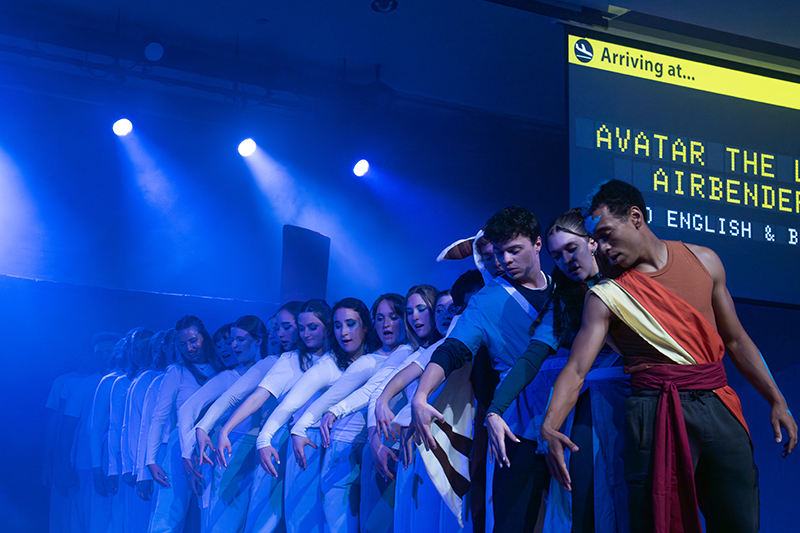 Stage performers in blue light doing a synchronized movement sequence, with a transit-style sign displaying “Avatar the Airbender.”
