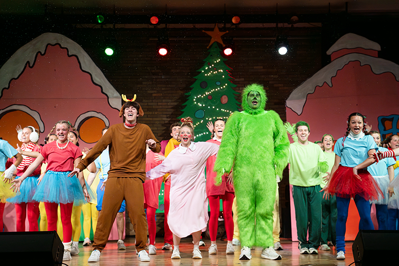 How the Grinch Stole Christmas team performing in colorful costumes, including the Grinch in a bright green suit in front of a decorated Christmas tree.