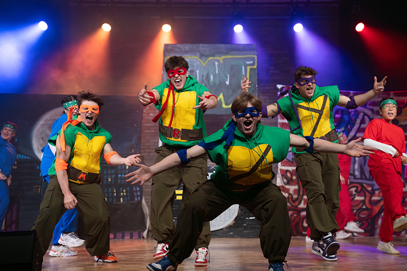 Performers in Teenage Mutant Ninja Turtles costumes dancing on a stage under colorful spotlights.