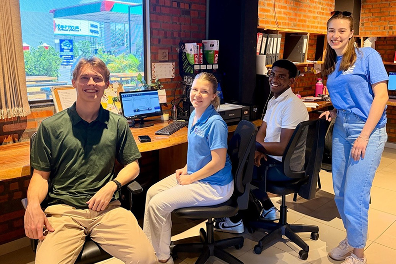 Four students smiling in a brick-walled office, seated and standing beside desks with computers near a large window.