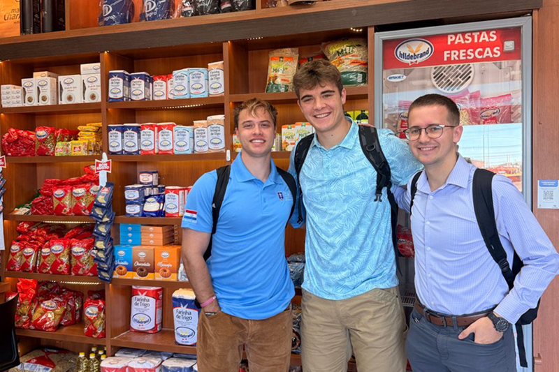 Three men with backpacks smile while posing in a pasta shop aisle, with shelves of packaged goods and a freezer labeled “Pastas Frescas” behind them.