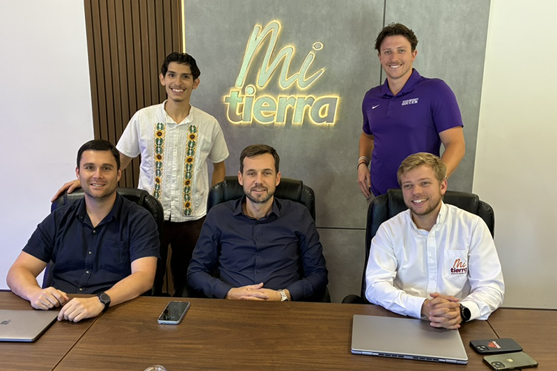 Five men pose around a conference table in an office, with a lit “Mi tierra” wall sign behind them.