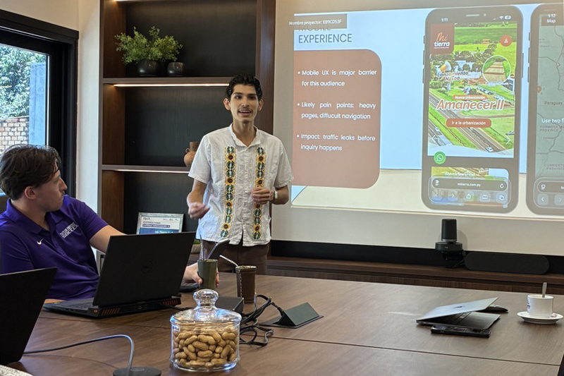 Person presenting in a conference room beside a projected slide about mobile UX, while colleagues sit at a table with laptops and meeting materials.