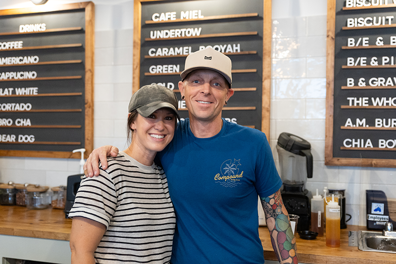 Bethany and Justin stand behind the counter with their arms around each other, with menu boards in the background.