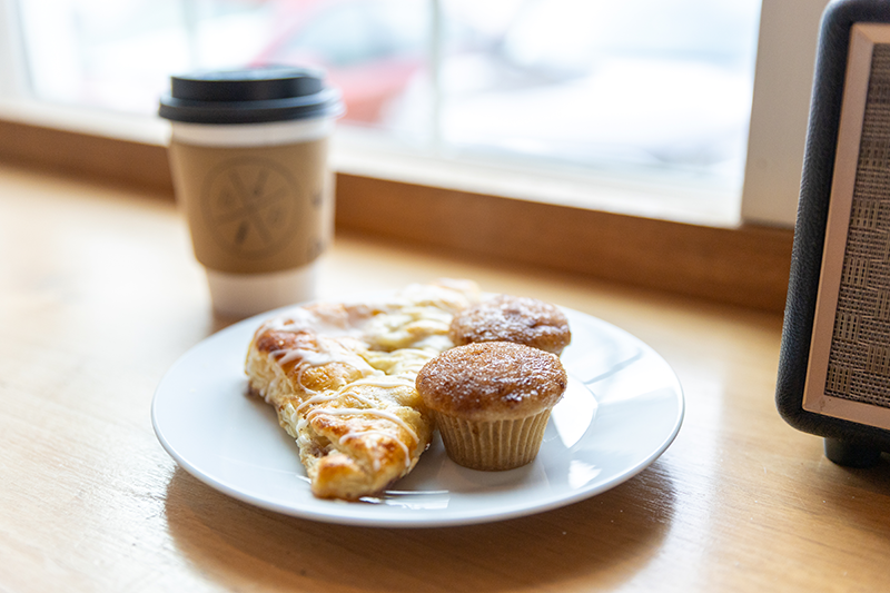 Plate with a slice of pizza and two muffins on a wooden table beside a takeaway coffee cup near a window.