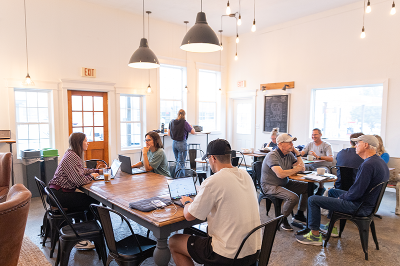 People working on laptops and chatting at shared tables in a bright, modern cafe with large windows and hanging pendant lights.