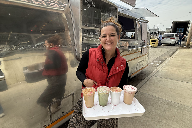 Avery smiling holding a tray of four colorful drinks in front of a shiny food truck.