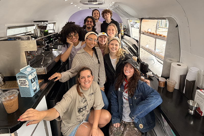 Smiling team of baristas gathered inside a coffee trailer, surrounded by an espresso machine, milk cartons, and ready-to-serve drinks.
