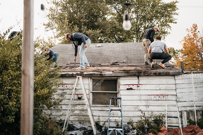 Several people repair a weathered roof on a small house, with ladders and tools set up below.