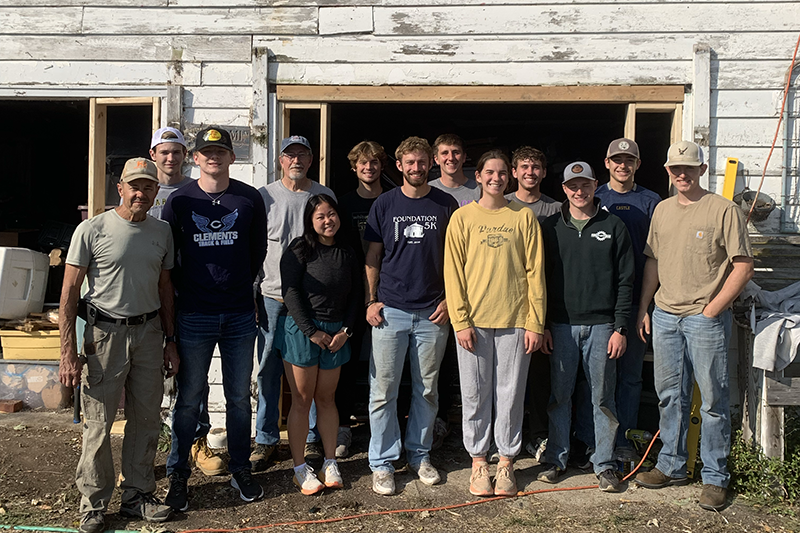 Group of volunteers standing in front of a weathered building with open doorways, during a work project.