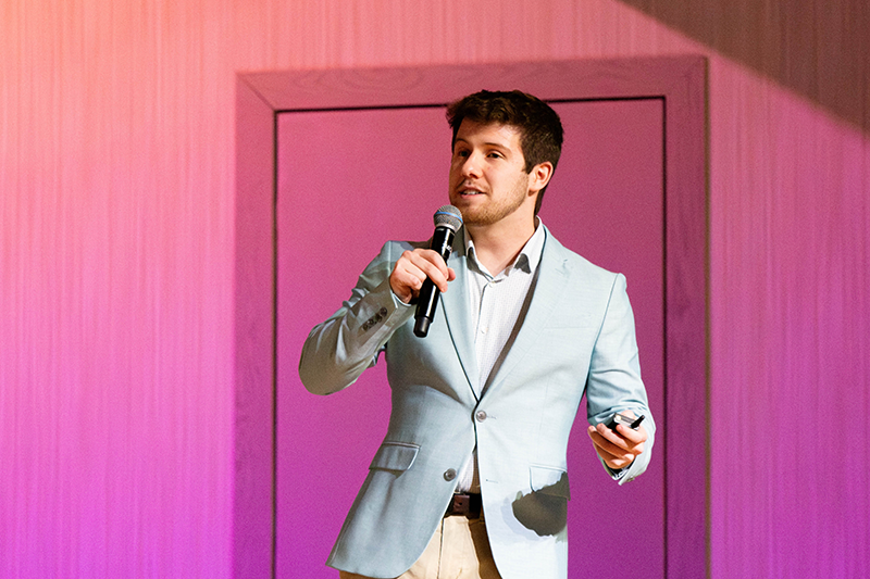 Andrew Beaverson in a light blue blazer speaking into a handheld microphone and holding a remote, standing in front of a purple backdrop.