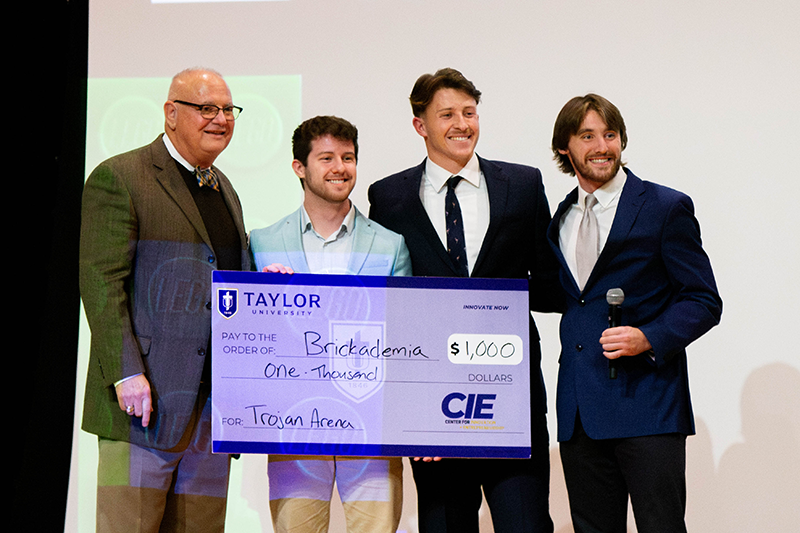 Four people on a stage hold a large ceremonial check from Taylor University for $1,000, while one person on the right holds a microphone.