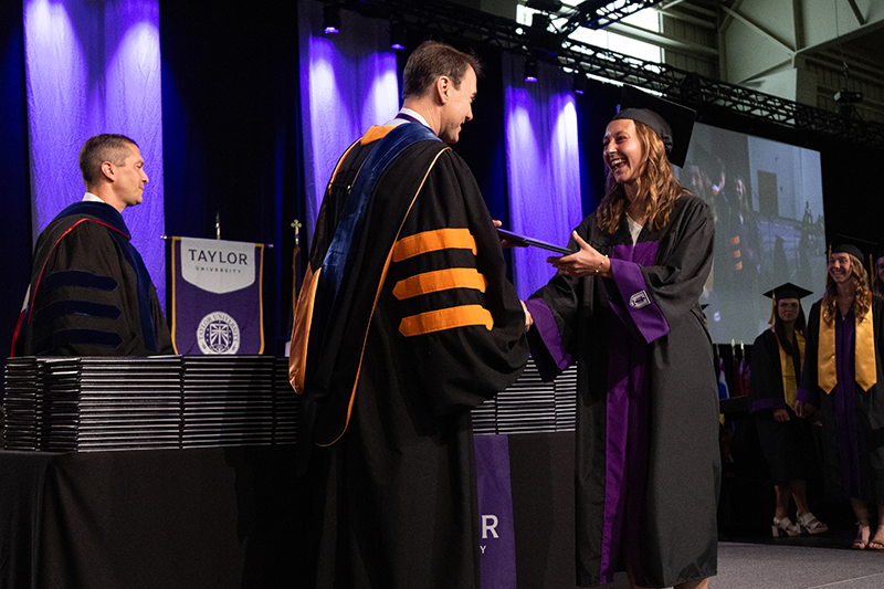 Graduate in cap and gown smiles while shaking hands with President Lindsay onstage at a Taylor University commencement ceremony.
