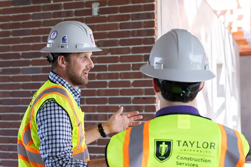 Two men in hard hats and reflective vests talk at an indoor site site, one gesturing while facing a brick wall.