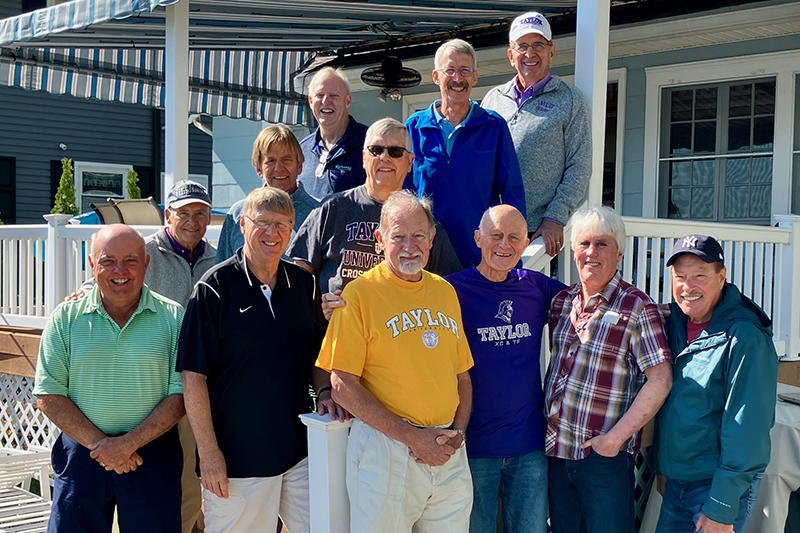 Group of men posing and smiling on a porch in front of a house with striped awnings.