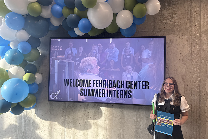Falon holding a booklet and a green folder stands beside a screen reading Welcome Fehribach Center Summer Interns under a blue, green, and white balloon arch.
