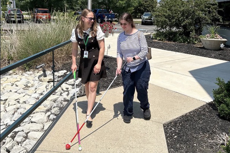 Falon and another woman walk along a sidewalk outdoors using white canes for navigation.