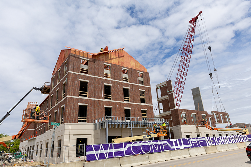 Multi-story brick building under construction with workers on lifts and a tall crane beside it, under a partly cloudy sky.