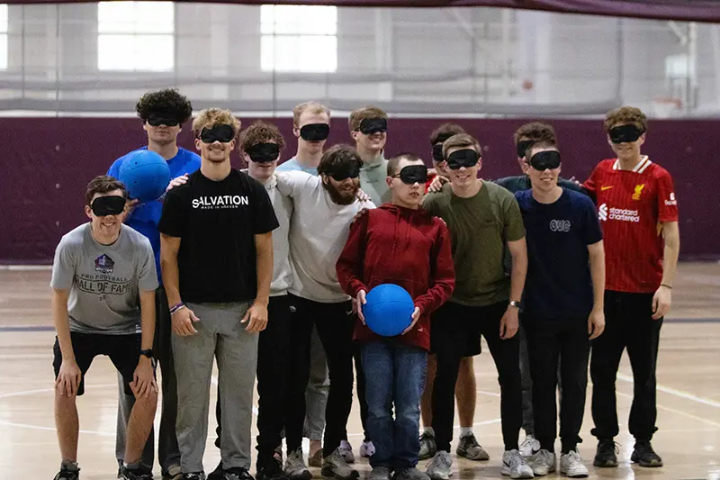 Group of thirteen students wearing eyeshades pose for a photo after playing goalball