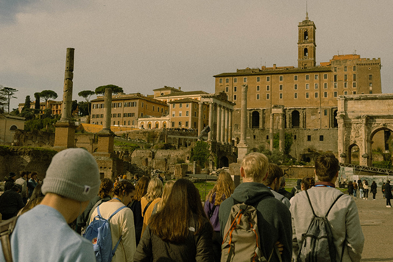 Students walking through ancient ruins toward a large historic building with a bell tower in the background.