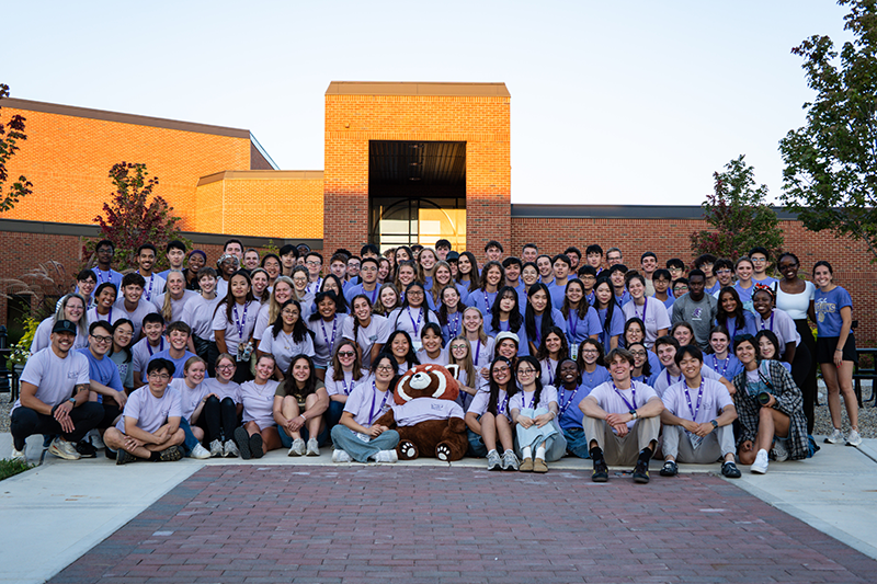 Large group of students and staff posing for a group photo in front of a brick school building, with a mascot plush in the front row.