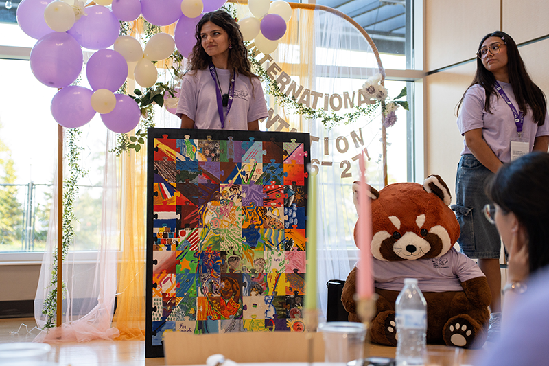 Two volunteers stand by a colorful collage artwork at a decorated event booth with purple-and-white balloons and a large stuffed red panda.
