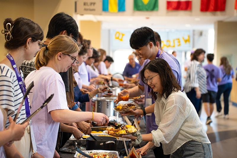 People line up at a buffet table as volunteers serve food and drinks in a busy hallway with flags hanging overhead.