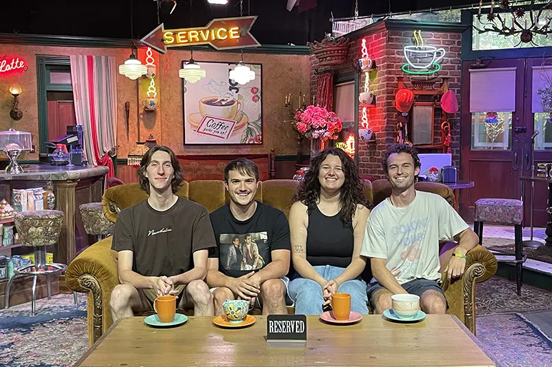 Four students sitting on the classic Friends couch at a café-themed set, with colorful mugs and a “Reserved” sign on the table.