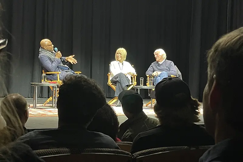 Panel discussion on a stage with three speakers seated in chairs, viewed from the audience.