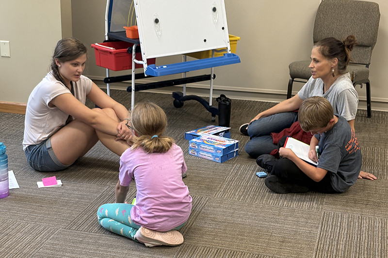 Small group session in a classroom-like room, with an adult and three children sitting on the floor while one child writes in a notebook.