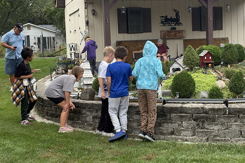 Children and adults gather around an outdoor model train display in a landscaped yard beside a house.