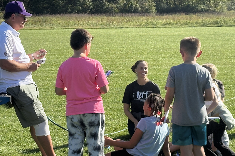 Adults and children gathered on a grassy field during an outdoor activity, with one adult holding papers and a seated woman smiling at the group.