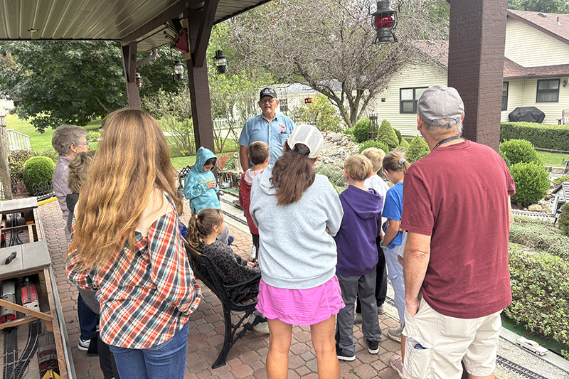 Group of children and adults gathered on a covered patio listening to a man speaking, with a landscaped yard in the background.