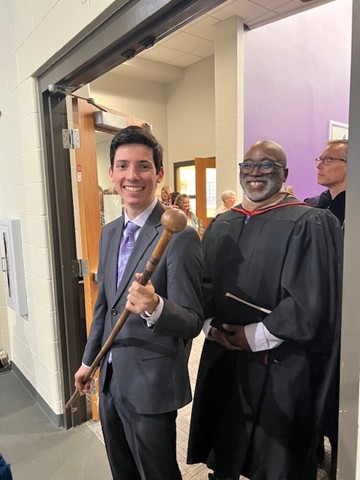 Smiling student in a suit holding a ceremonial mace beside a faculty member in academic robes backstage.