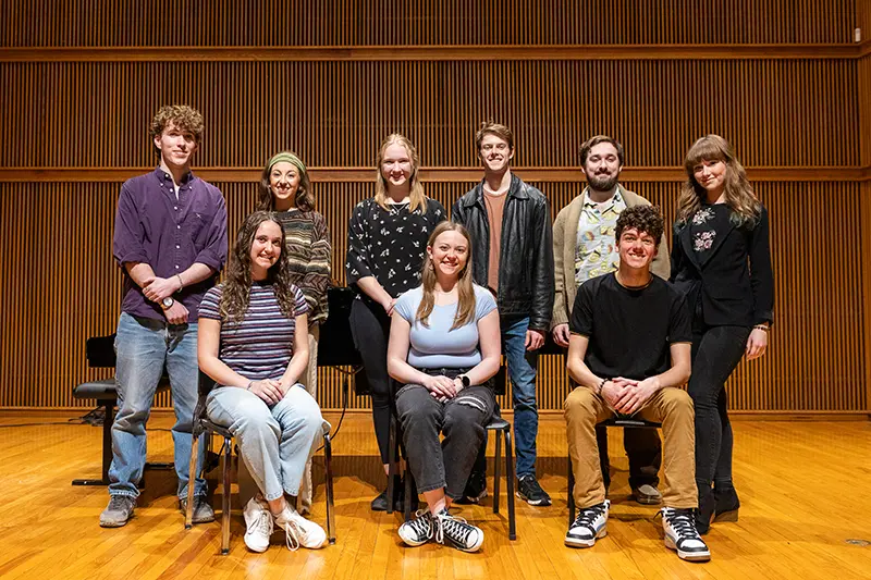 Nine students pose for a group photo on an auditorium stage, with two seated in front and the rest standing behind against a wood-paneled backdrop.