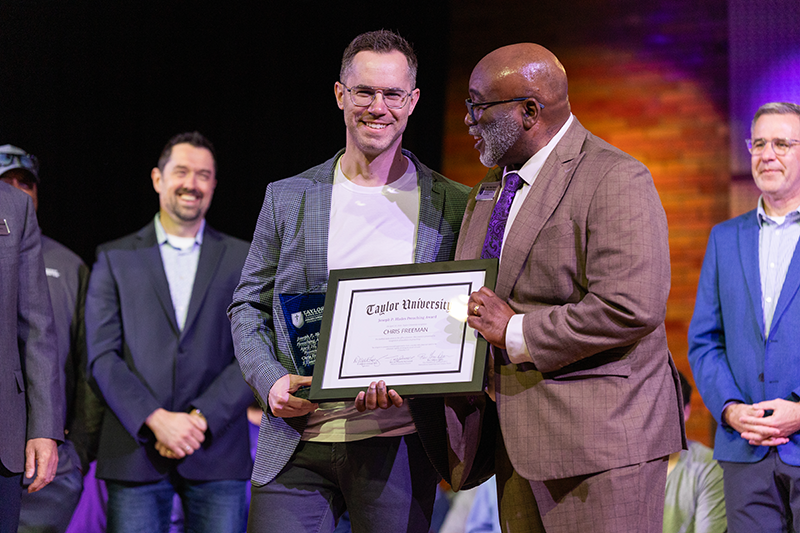 Two men on stage smile while holding a framed Taylor University certificate during an award presentation, with other attendees in the background.