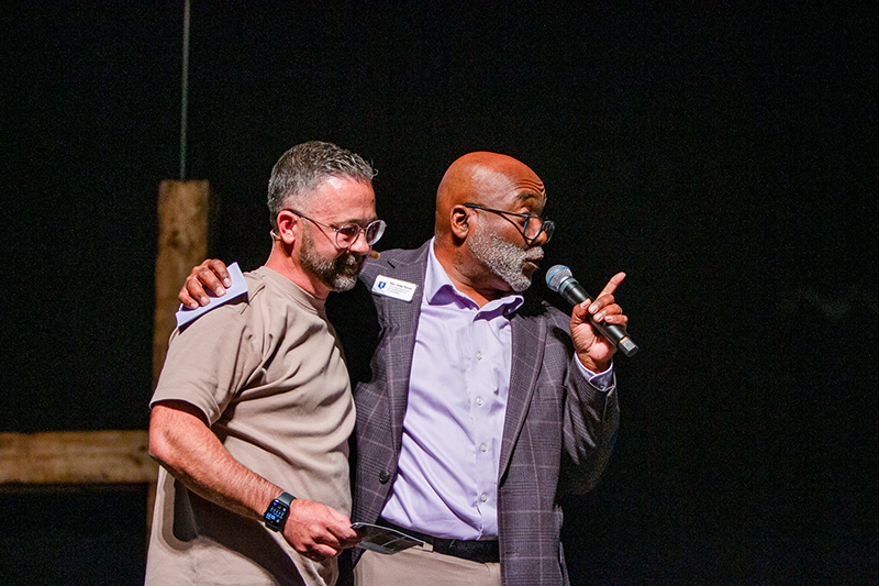Greg and Paul Maurer; one holds a microphone and gestures while the other stands beside him with an arm around his shoulder.